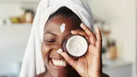 A woman wearing a towel on her head and smiling as she holds up a jar of cream in front of her eye. She has a strip of moisturizer on her forehead and on her nose.