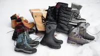 A number of pairs of winter boots, from various brands, laid out on a bed of snow.