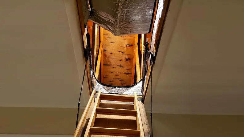 Standing at the bottom of an attic stairway, looking up to see the opening covered by an attic insulator.