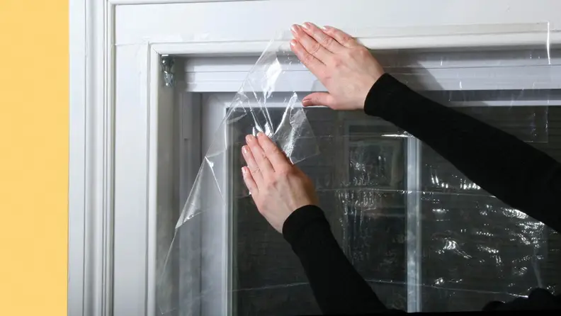 A close-up of someone affixing a layer of plastic insulation over a window.
