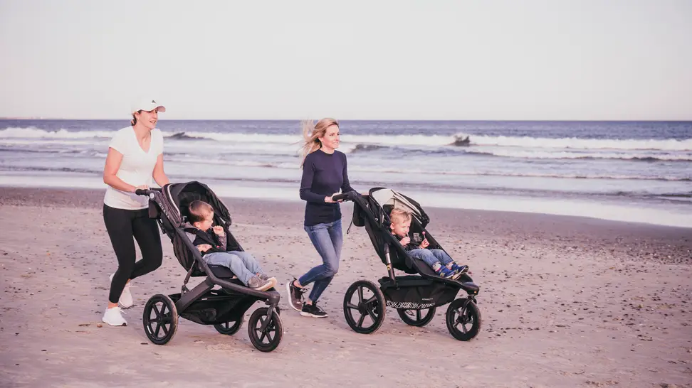 Two women jogging on a beach pushing kids in jogging strollers