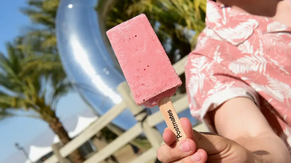 A person in a pink floral shirt holds a pink popsicle close to the camera lens.