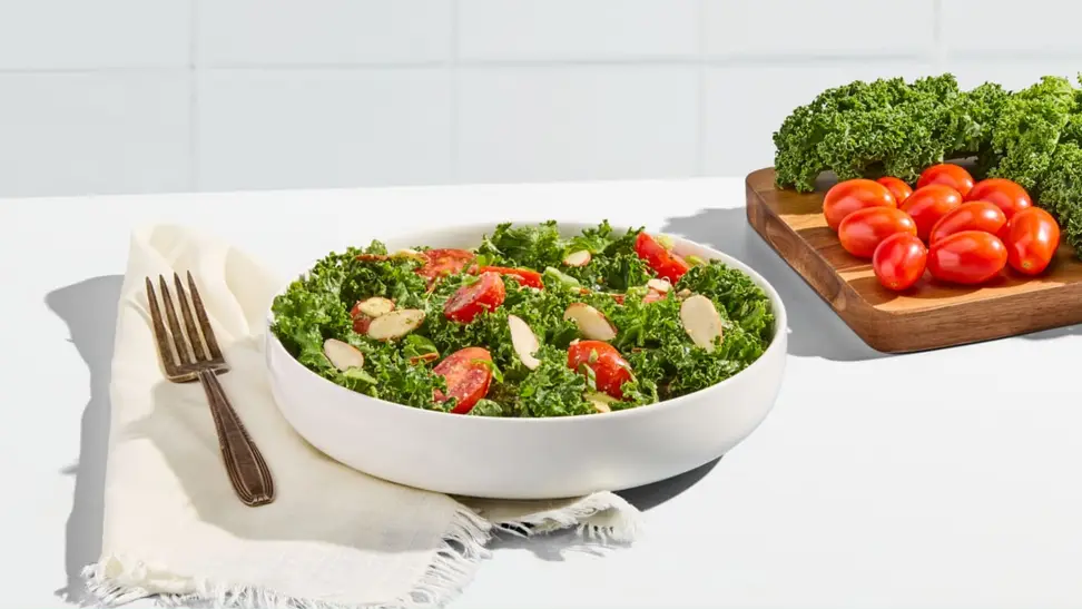 A bowl of salad on a white background next to a cutting board with kale and tomatoes.